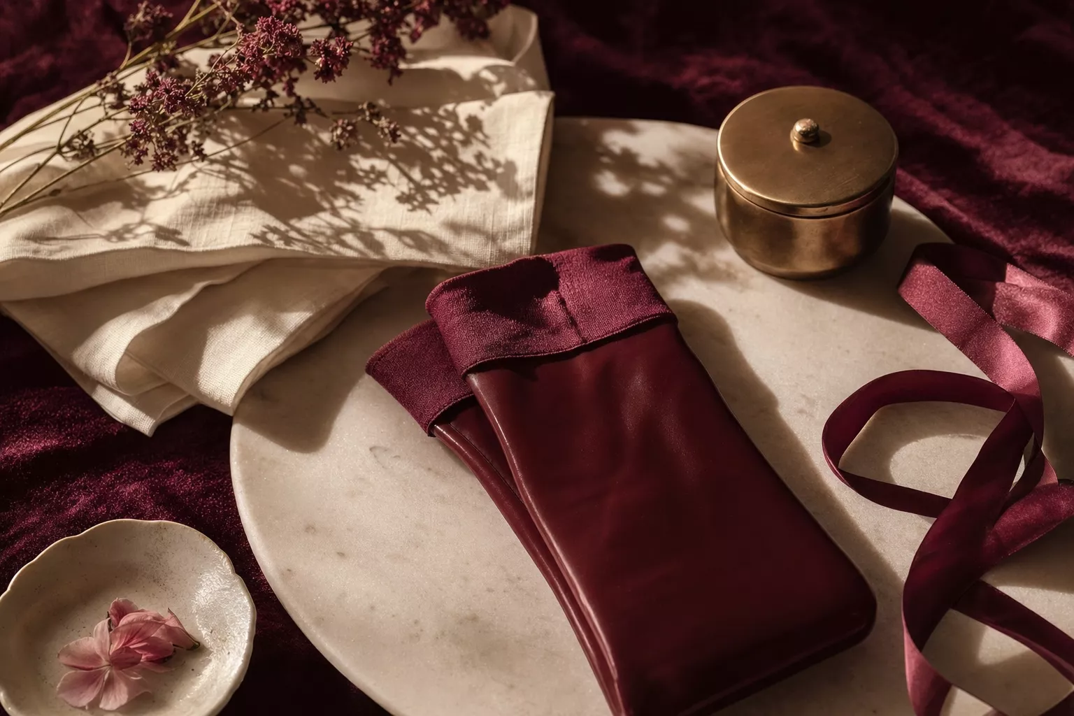 Nature morte éditoriale : bas vinyl pliés sur marbre avec ruban en soie bordeaux, textures ivoire, éclairage chaud.